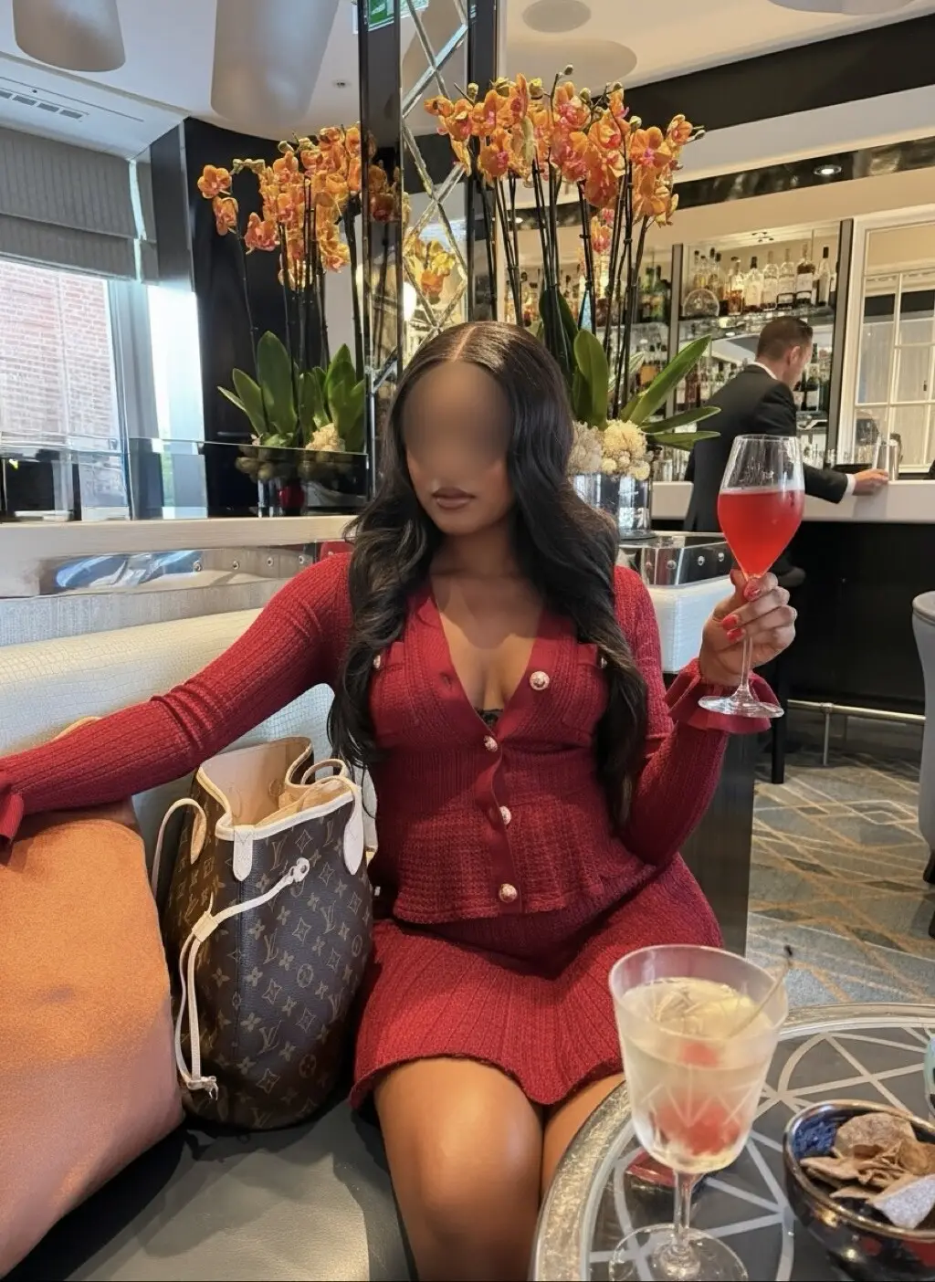 Woman in a red knit cardigan and skirt seated with a pink cocktail beside a second drink.