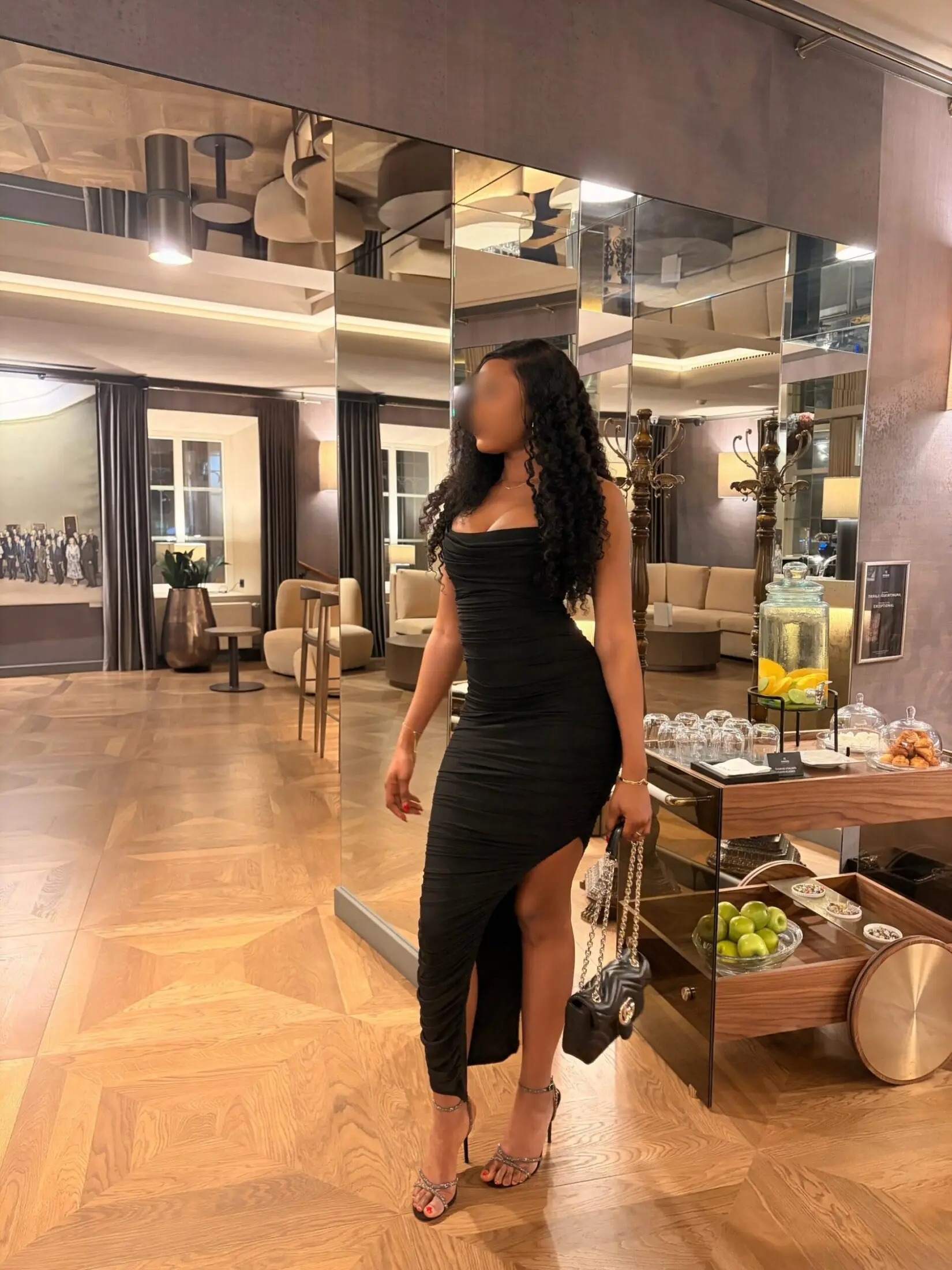 Woman in a black ruched dress with a high slit, standing on parquet and holding a handbag.