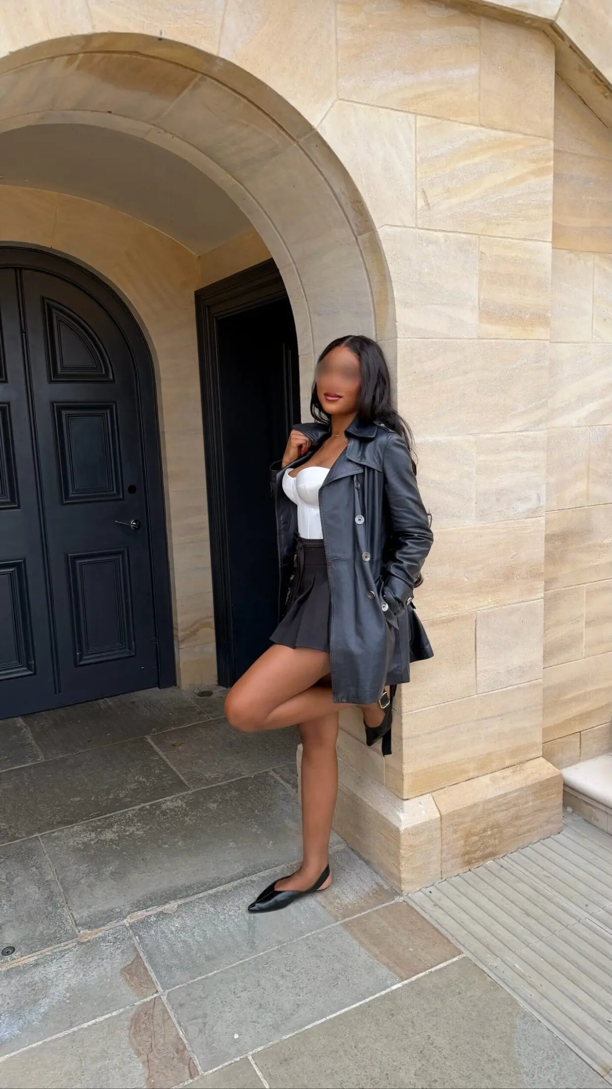 Woman in a black leather coat and mini skirt leaning on a sandstone wall beside a dark door.