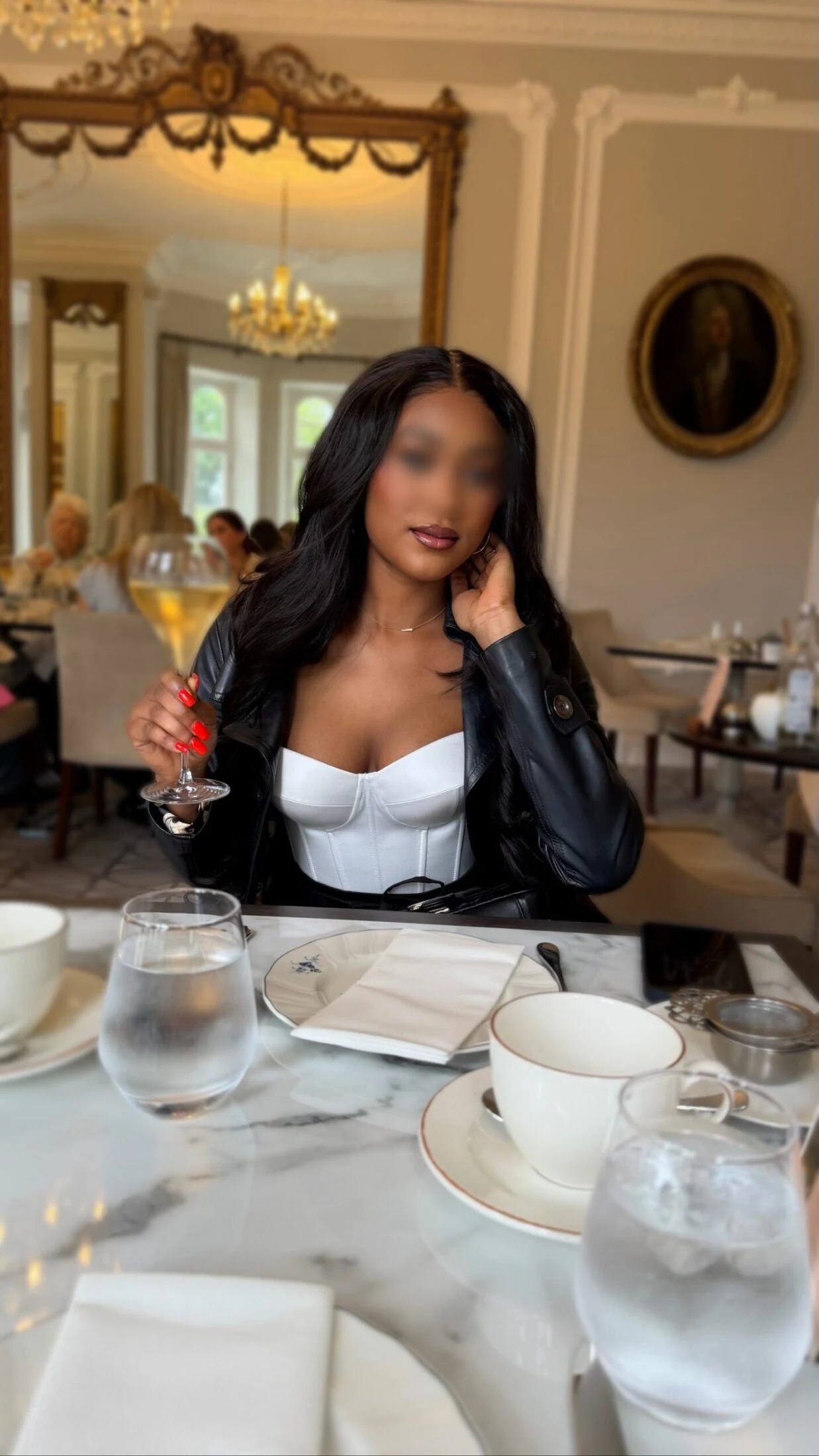 Woman in a leather jacket and white corset top seated at a marble table with a wine glass.