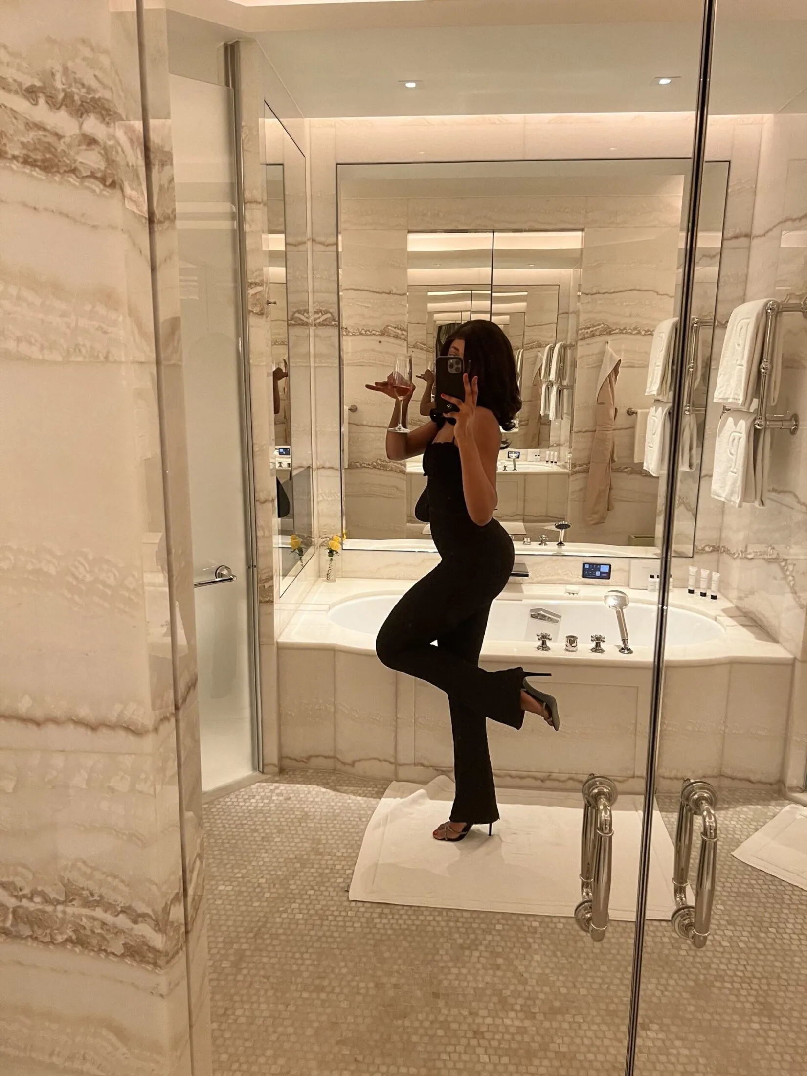 Woman in a black strapless jumpsuit holding a wine glass and phone in a marble bathroom.