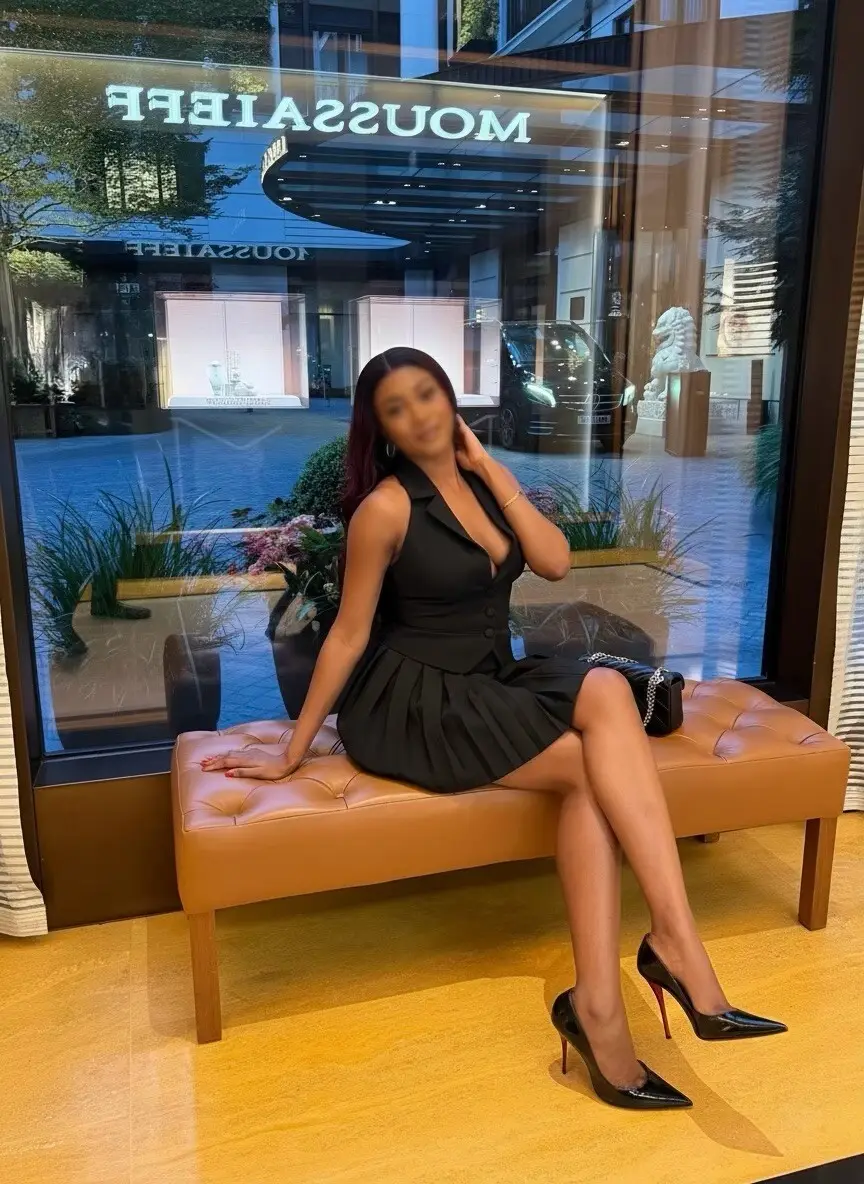 Woman in a black sleeveless pleated dress seated on a tan tufted bench with crossed legs.