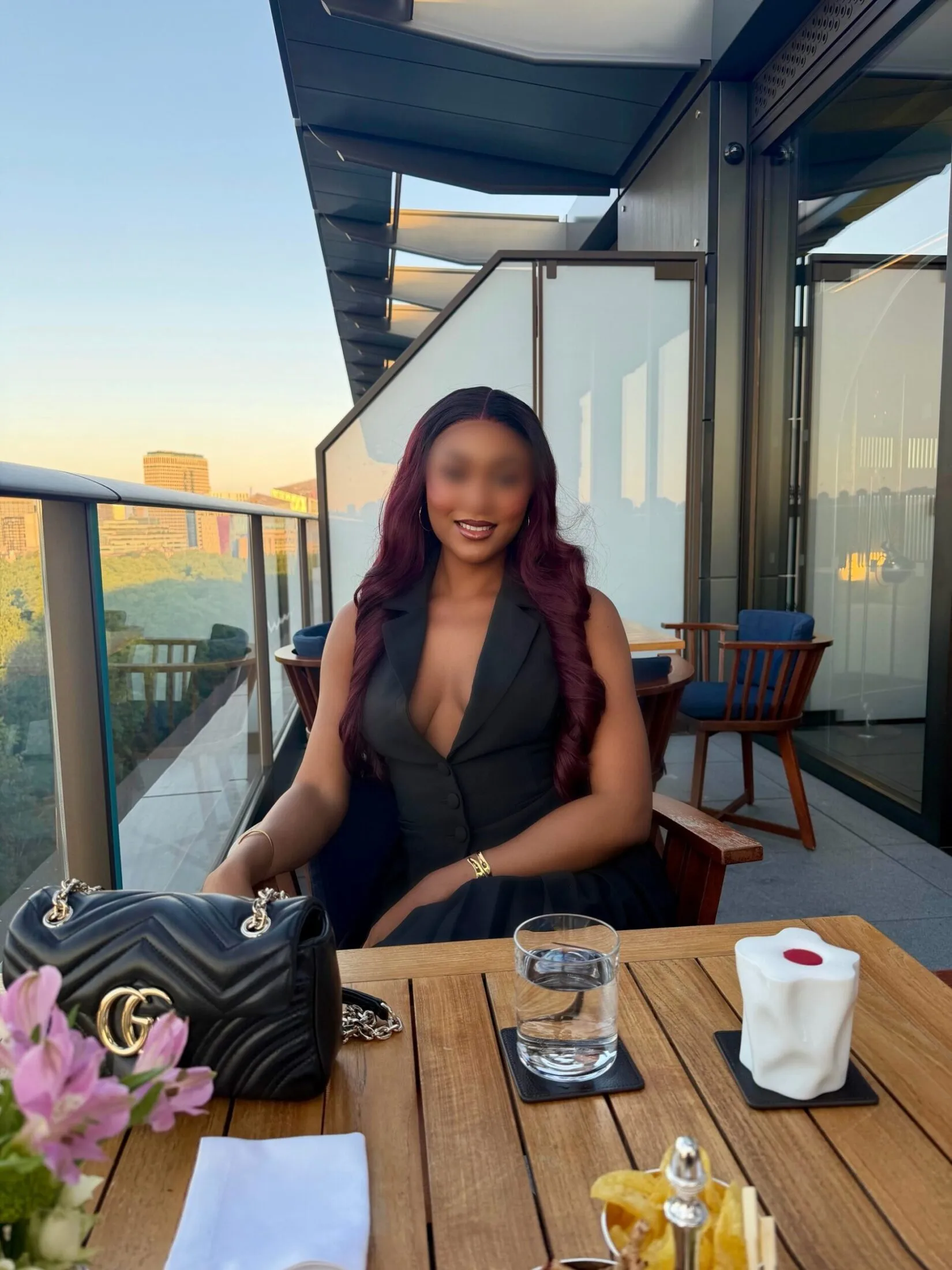 Woman in a black sleeveless blazer dress seated at a wooden table on an outdoor terrace.