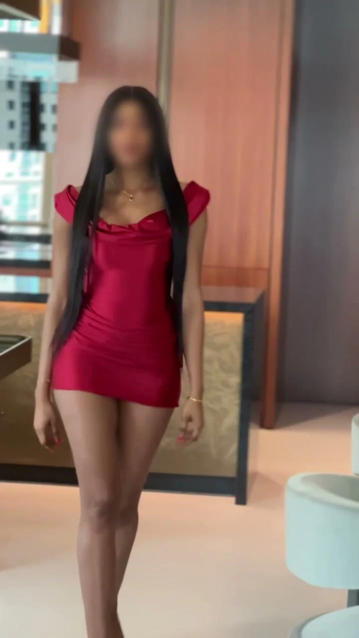 Film of a woman in a red satin mini dress walking towards the camera, then seated on a white sofa.